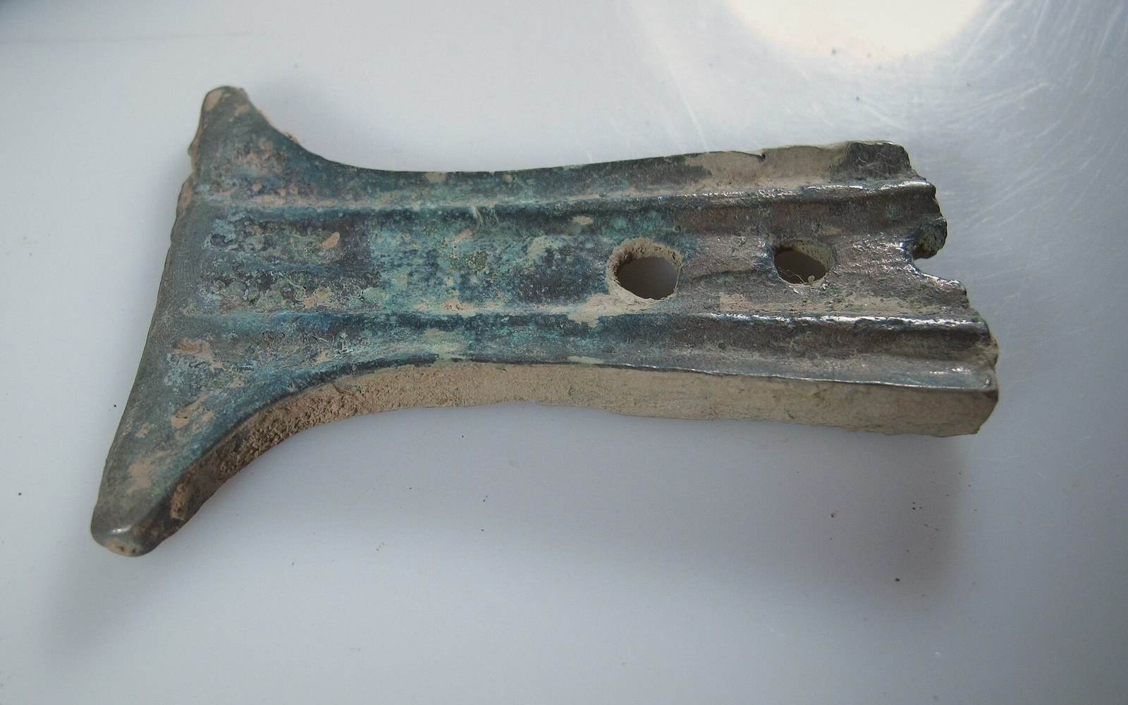 A corroded bronze axe head with two drilled holes lying on a white surface