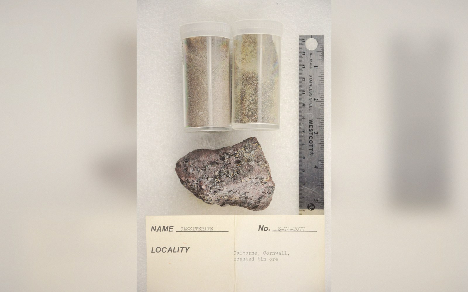 Cassiterite ore sample and two vials of roasted tin ore beside a ruler and label card