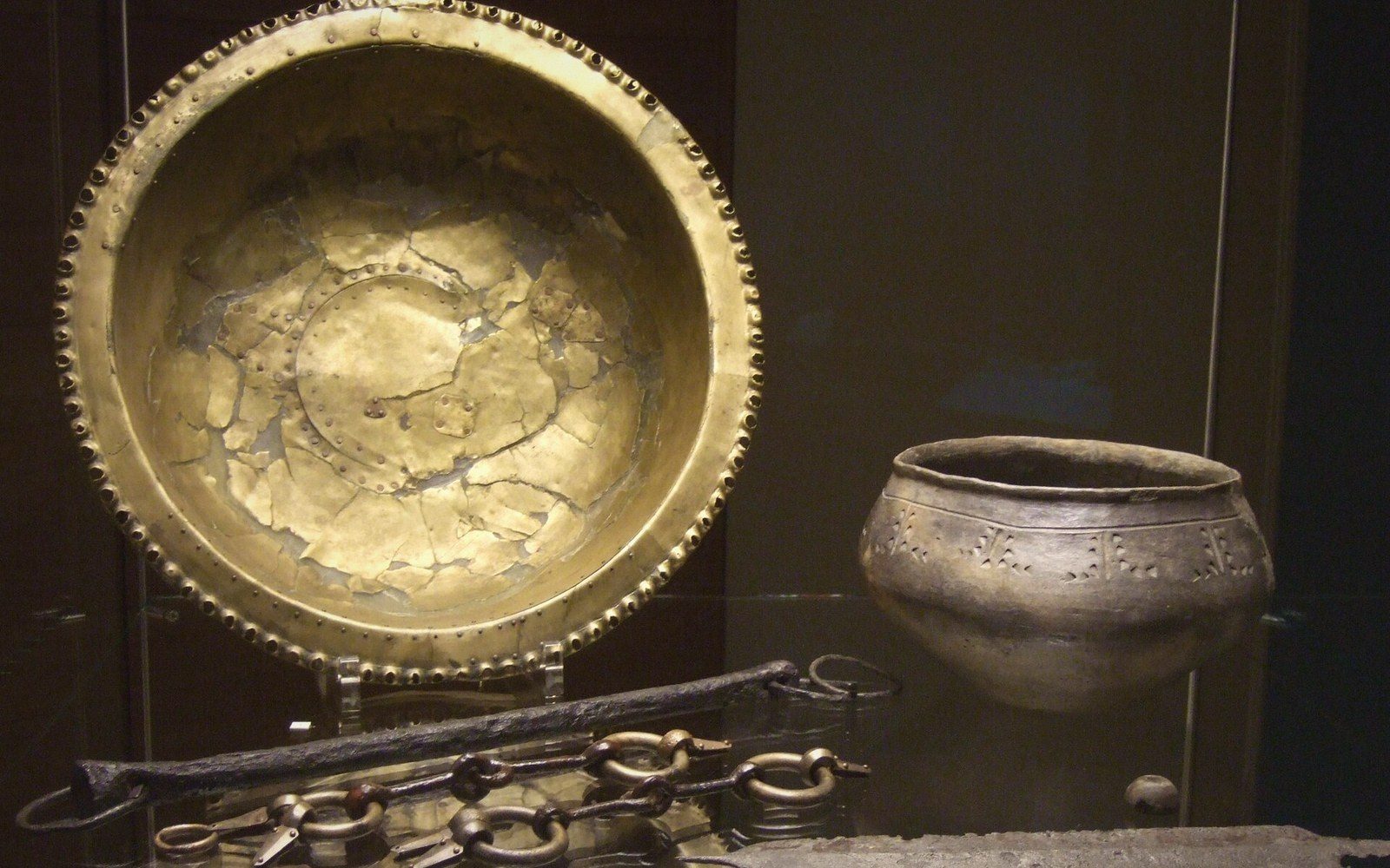 Ancient gold bowl, ceramic pot, and iron chain artifacts displayed together in a museum case
