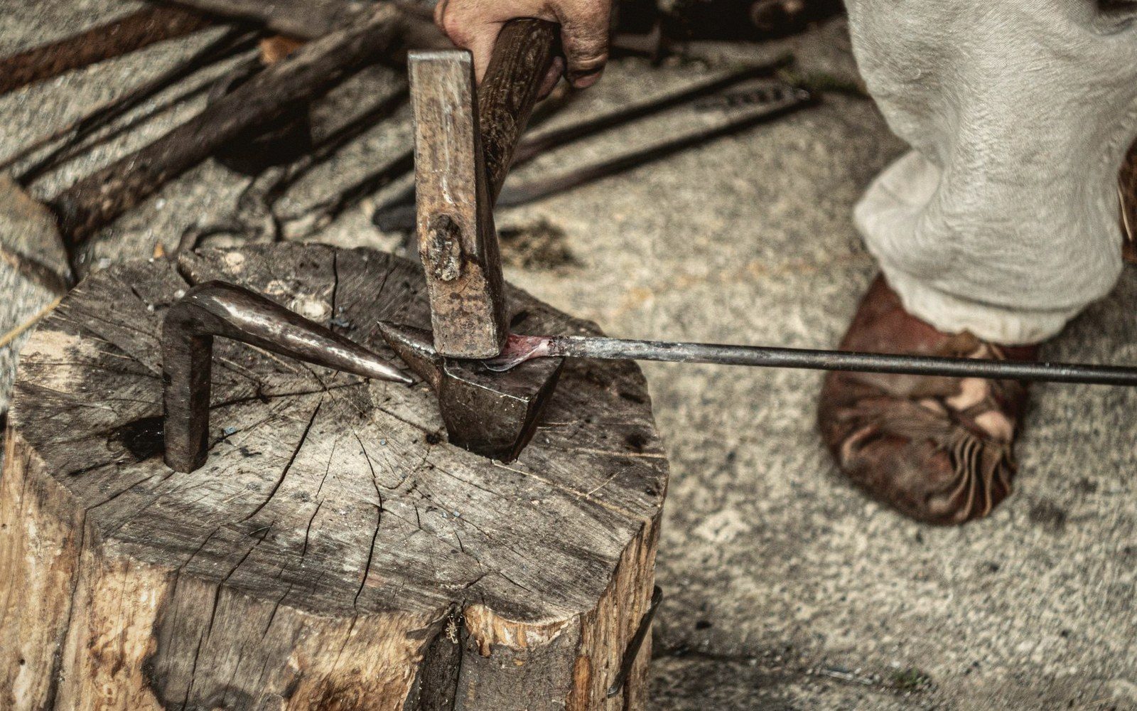 A blacksmith hammers a heated metal blade against a small anvil on a wooden block